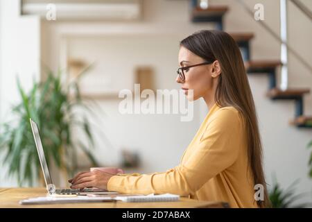 Focused millennial woman in glasses sitting on table, remote work on laptop, analyzing project statistics information, replies to customer emails from Stock Photo
