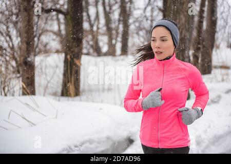 Winter sport fitness girl running in snow wearing wind jacket, gloves, headband and smartwatch. Asian woman healthy and active lifestyle training Stock Photo