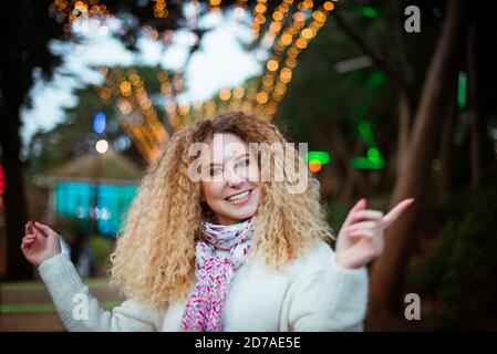 Portrait of Christmas happy smiling blonde curly woman wearing knitted white sweater and dancing or singing on decorated park background. Celebrating Stock Photo