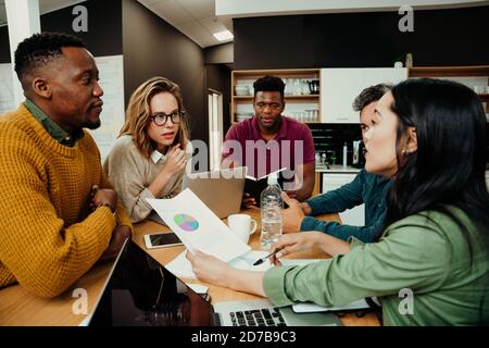 Diverse team gathered during lunch breaks working on laptop researching through documents discussing work Stock Photo