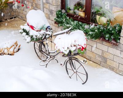 A metal decorative bicycle covered with snow stands in front of a window on the street during a snowfall Stock Photo