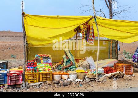 Woman stallholder sitting cross-legged in a stall selling spices and local goods in a roadside market in a village in Madhya Pradesh, India Stock Photo