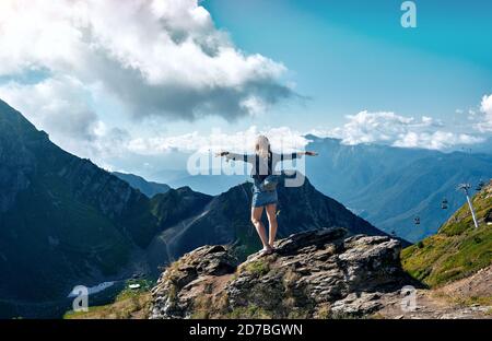 Young woman standing with raised hands with backpack on cliff's edge and looking into a wide valley Stock Photo