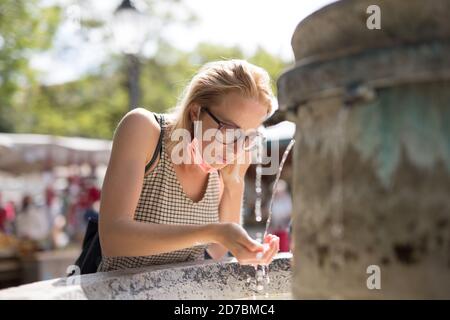 Thirsty young casual cucasian woman wearing medical face mask drinking water from public city fountain on a hot summer day. New social norms during