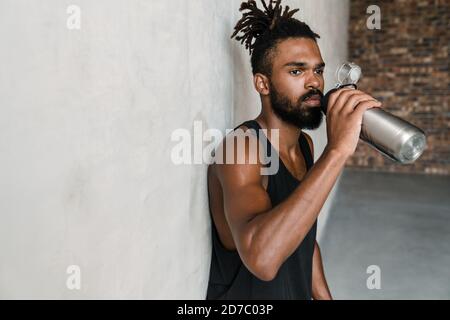 Image of handsome african american sportsman drinking water while leaning on wall indoors Stock Photo