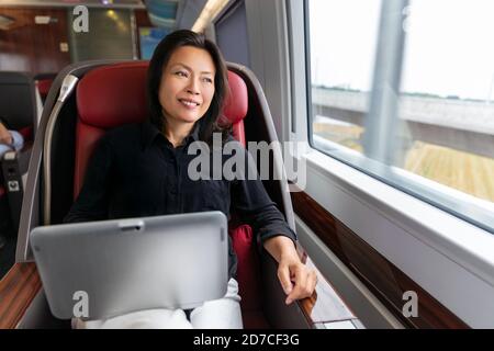 Business trip Asian woman working on laptop on train commute travel lifestyle. Middle aged chinese businesswoman smiling looking out window in