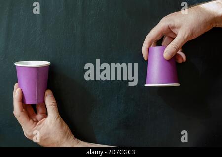 Creative background with paper cups for hot drinks and a man's hand. Hand and lilac glasses for coffee on a black background. Top view at an angle. Stock Photo