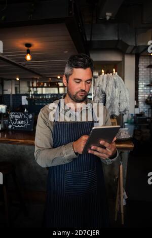 Male waiter scrolling through menu on digital tablet wearing apron in trendy cafe  Stock Photo