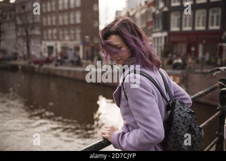 Girl in the coat and backpack enjoying Amsterdam city. Young woman looking to the side on Amsterdam channel, Netherlands, Europe. Stock Photo