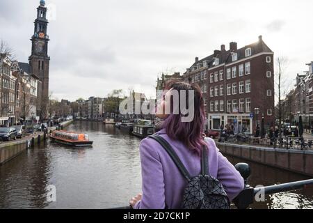 Girl in the coat and backpack enjoying Amsterdam city. Young woman looking to the side on Amsterdam channel, Netherlands, Europe. Stock Photo