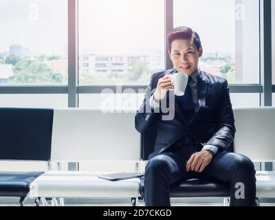 Happy Asian businessman in suit drinking white cup of hot coffee while sitting on waiting chair near huge glass window with sunlight with copy space. Stock Photo