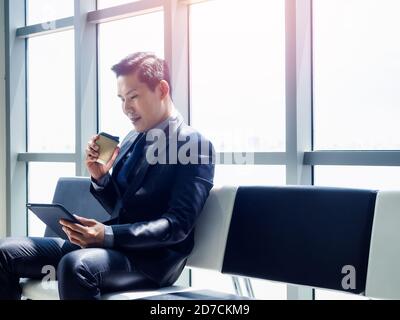Good looking Asian businessman in suit watching tablet on his hand and holding disposable paper coffee cup while sitting on waiting chair near huge gl Stock Photo