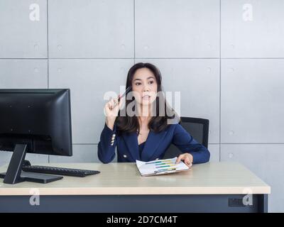 Asian business woman long hair in blue suit thinking and working with desktop computer in office on grey wall background. Beautiful secretary holding Stock Photo