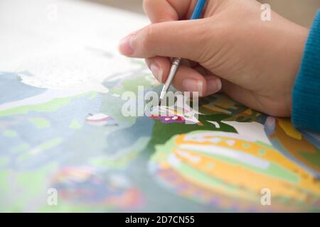 1 - Close up of young female hand painting by numbers. Blue colors of jumper cuff and brush handle pop against the warm skin tones. Selective focus Stock Photo