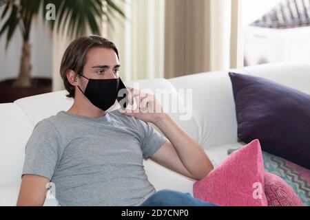 Close up of a young man using a face mask sitting on a sofa and talking on his phone on an out of focus background. Communication and safety concept. Stock Photo