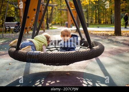 two scared toddlers boy and girl left unattended are sitting on a swing hammock on a playground in the autumn park Stock Photo