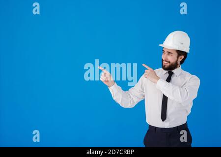 Handsome man industrial engineer wearing a white hard helmet and pointing finger towards something blank copy space for text isolated on blue Stock Photo