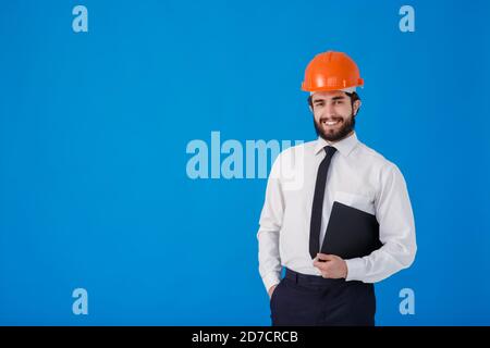 A young bearded male construction worker in a white shirt and construction helmet on a blue background. industrial engineer-Architect hold clipboard Stock Photo