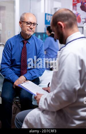 Old man answering doctor questionnaire during examination in hospital room. Converstation with medical stuff clinic medicine healthcare, senior, coat, clipboard. Stock Photo