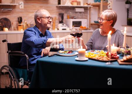 Disabled man smiling at wife while having dinner in kitchen. Wheelchair immobilized paralyzed handicapped man dining with wife at home, enjoying the meal. Stock Photo