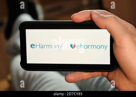 Eharmony logo on the screen of mobile phone in mans hand. Application for dating and acquaintance. Man is laying on the sofa and looking for partner Stock Photo