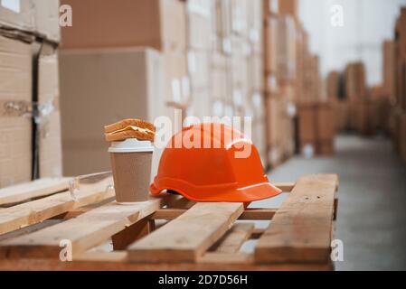 Close up view of orange colored hard hat, cup of drink and sandwich in the warehouse Stock Photo