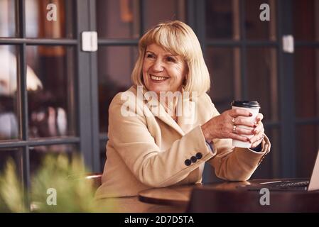 Portrait of senior woman that sits indoors in the cafe with modern laptop and cup of drink in hands Stock Photo