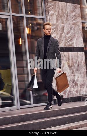 Man in elegant formal wear and with shopping packages is outside against modern building Stock Photo