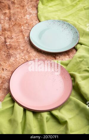 Two empty blue and pink plates on brown concrete background and green textile. Side view, close up. Stock Photo