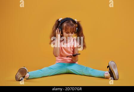 Portrait of cute little girl in headphones and with phone in the studio against yellow background Stock Photo