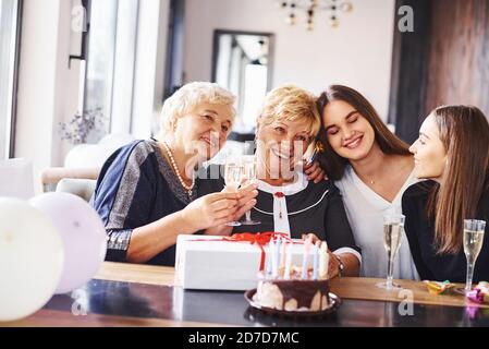 Glasses with alcohol in hands and cake on table. Senior woman with family and friends celebrating a birthday indoors Stock Photo