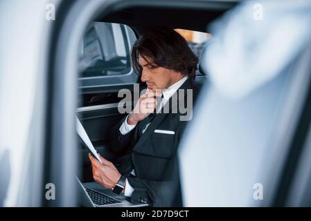 Using notepad. Young businessman in black suit and tie inside modern automobile Stock Photo
