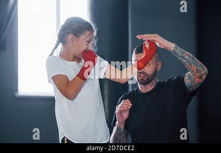 Basic punches. Young tattooed boxing coach teaches cute little girl in the gym Stock Photo