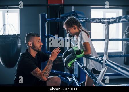 Conception of assistance. Young tattooed boxing coach teaches cute little girl in the gym Stock Photo