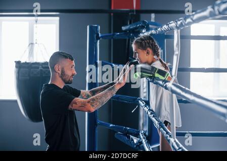 Conception of assistance. Young tattooed boxing coach teaches cute little girl in the gym Stock Photo