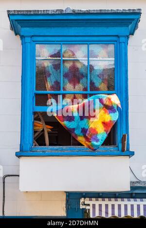 A brightly coloured curtain blowing from an old blue upstairs window left open on a windy day, UK Stock Photo