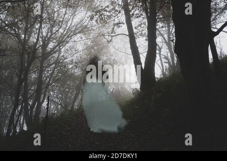 A spooky ghostly woman in a long white dress walking up a path through a moody misty autumn woodland Stock Photo