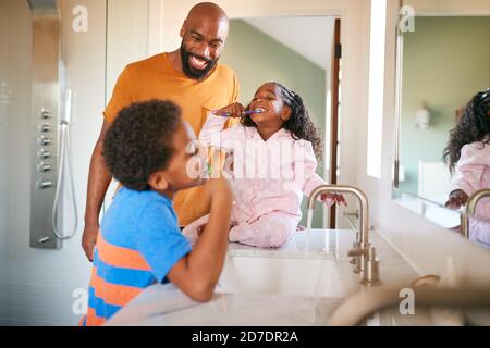 Father Helping Children To Brush Teeth In Bathroom At Home Stock Photo