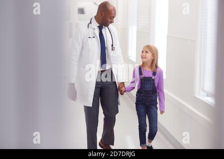 Male Paediatric Doctor Walking Along Hospital Corridor Holding Hands With Young Girl Patient Stock Photo