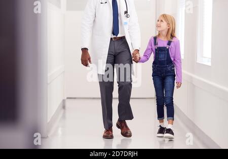 Close Up Of Male Paediatric Doctor Walking Along Hospital Corridor Holding Hands With Girl Patient Stock Photo