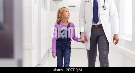 Close Up Of Male Paediatric Doctor Walking Along Hospital Corridor Holding Hands With Girl Patient Stock Photo