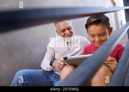 Grandfather Sitting On Steps Outdoors At Home With Grandson Using Digital Tablet Stock Photo