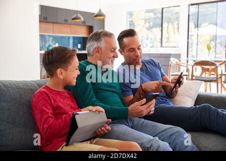 Multi-Generation Male Hispanic Family Sitting On Sofa At Home Using Mobile Phones And Digital Tablet Stock Photo