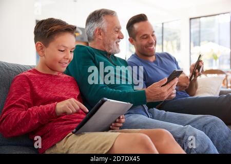 Multi-Generation Male Hispanic Family Sitting On Sofa At Home Using Mobile Phones And Digital Tablet Stock Photo