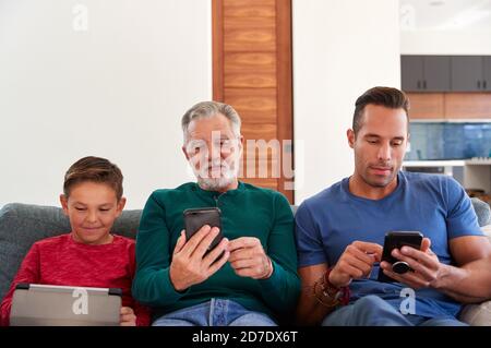 Multi-Generation Male Hispanic Family Sitting On Sofa At Home Using Mobile Phones And Digital Tablet Stock Photo