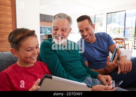 Multi-Generation Male Hispanic Family Sitting On Sofa At Home Using Mobile Phones And Digital Tablet Stock Photo