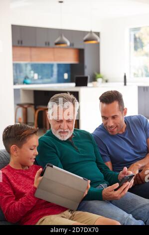 Multi-Generation Male Hispanic Family Sitting On Sofa At Home Using Mobile Phones And Digital Tablet Stock Photo