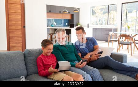 Multi-Generation Male Hispanic Family Sitting On Sofa At Home Using Mobile Phones And Digital Tablet Stock Photo