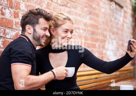 Young Couple On Date Taking Selfie On Mobile Phone Sitting At Outdoor Coffee Shop Table Together Stock Photo