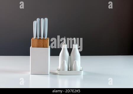 set of white porcelain vinegar, oil, salt and pepper shakers and kitchen knife set in white holder on the white table, close-up Stock Photo